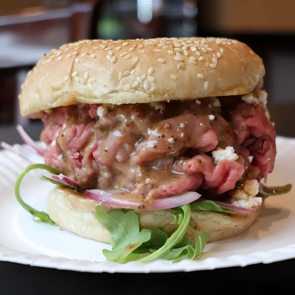 A roast beef sandwich with sauce, feta, red onion, and arugula on a sesame seed bun.