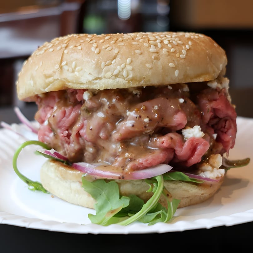 A roast beef sandwich with sauce, feta, red onion, and arugula on a sesame seed bun.