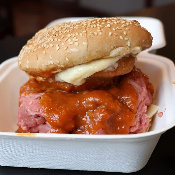 A close-up of a roast beef sandwich with red sauce and cheese in a white takeout container.